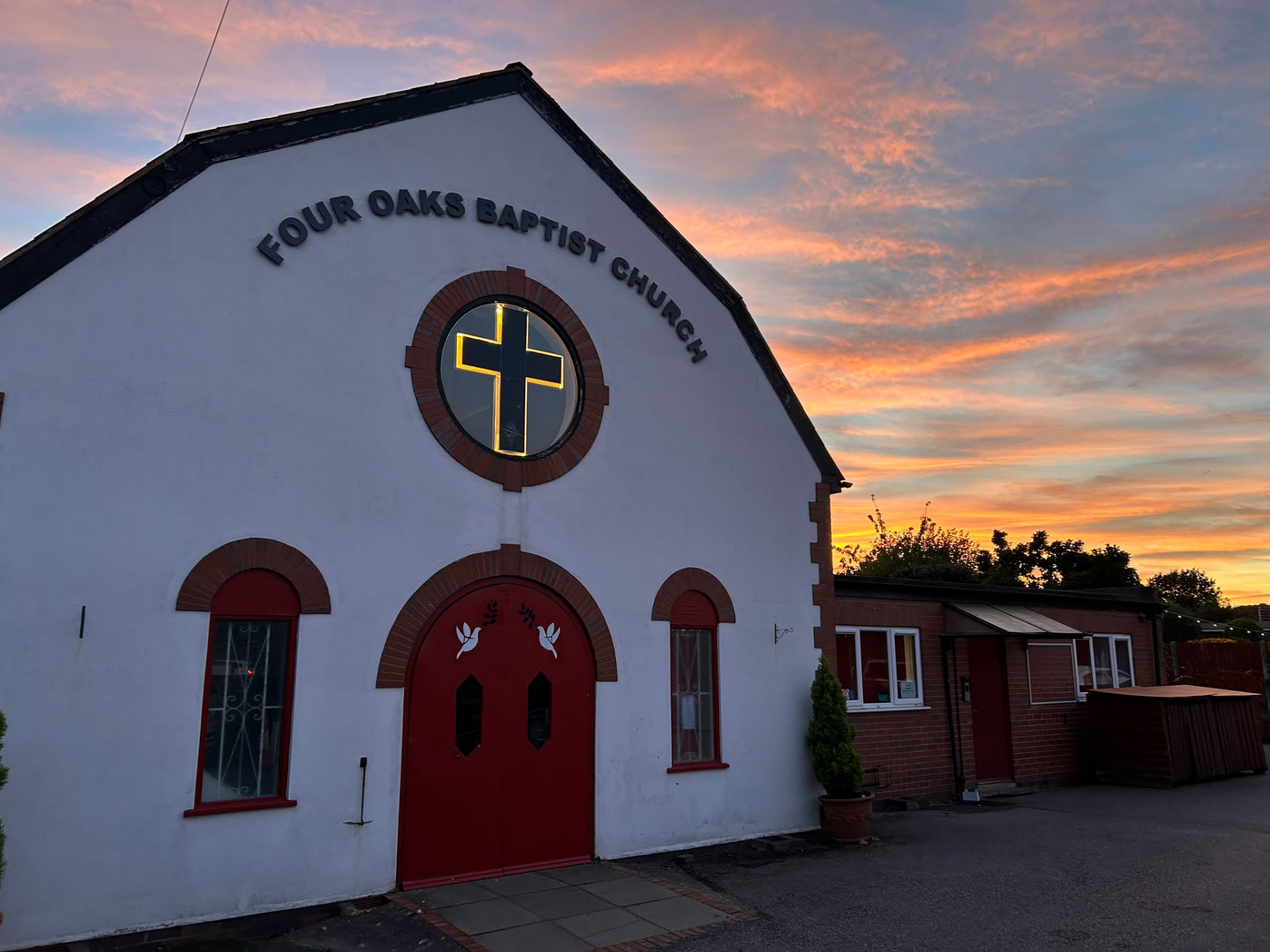 Four Oaks Baptist Church at sunset, glowing cross window under a pink and orange sky